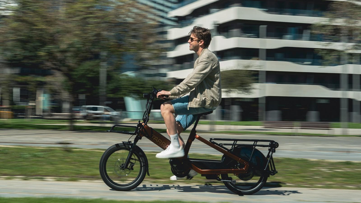 Mann fährt in der Stadt ein braunes Tarra-Elektro-Lastenrad mit Frontkorb und Gepäckträger hinten